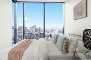 A modern bedroom with a large window offering a city view, a bed with white linens, and a black woven chair.
