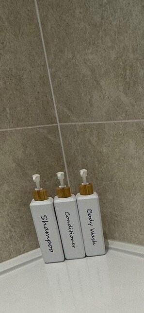 Tres botellas de productos de baño etiquetadas como 'Champú', 'Acondicionador' y 'Gel de ducha' sobre una superficie blanca frente a una pared con baldosas.