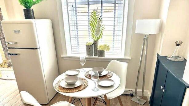 Un comedor con una mesa preparada para dos personas, una silla blanca, un refrigerador pequeño y una ventana con persianas y plantas.