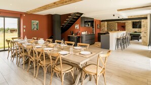 Une salle à manger spacieuse, dotée de chaises en bois et d’une longue table, une cuisine aux murs en pierre et un escalier menant à un niveau supérieur.