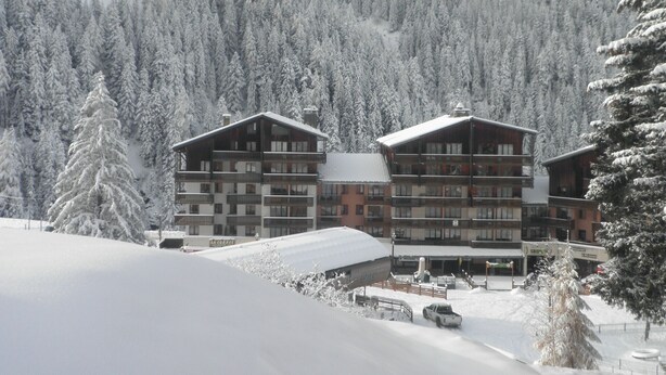 A multi-story hotel with balconies, surrounded by snow-covered trees and a mountainous backdrop.