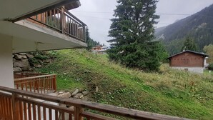 Un balcon doté d’un mur en pierre et d’une balustrade en bois, offrant une vue sur une région montagneuse, caractérisée par une végétation luxuriante et la présence d’un chalet en bois.