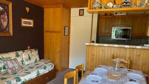 A cozy room with a wooden sofa, a dining table set for two, and a kitchen area with a refrigerator and a stove.