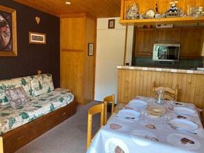 A cozy room with a wooden sofa, a dining table set for two, and a kitchen area with a refrigerator and a stove.
