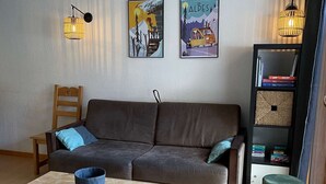Un saló tranquil i acollidor amb un sofà marró, una taula de centre de fusta i un petit tamboret. Hi ha dues làmpades de paret i una biblioteca amb llibres i objectes decoratius.