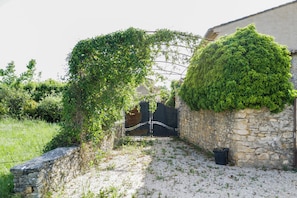 Stroll through the serene garden, surrounded by lush greenery and a rustic stone wall, to a charming building with a sloped roof.