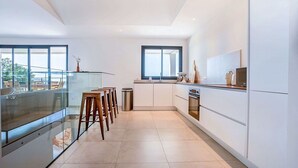 A modern kitchen with a glass railing, wooden stools, a large window, and a view of the outdoors. - Superb 6 Bedroom Villa, Sea View, Swimming Pool & Pétanque, 5mn from the Beach (Lumio)