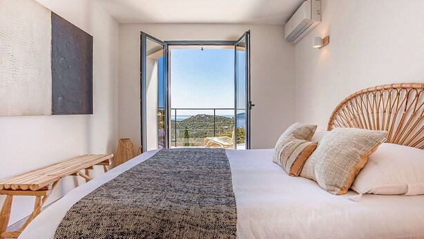 A bedroom with a bed, a wooden bench, a view of the outdoors, and a wall-mounted air conditioner. - Superb 6 Bedroom Villa, Sea View, Swimming Pool & Pétanque, 5mn from the Beach (Lumio)