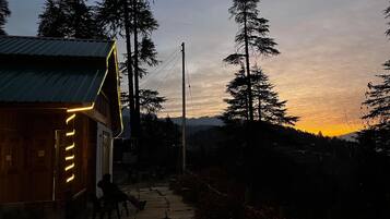 A wooden cabin with a porch and a person sitting on a bench. There are trees and a mountain in the background.