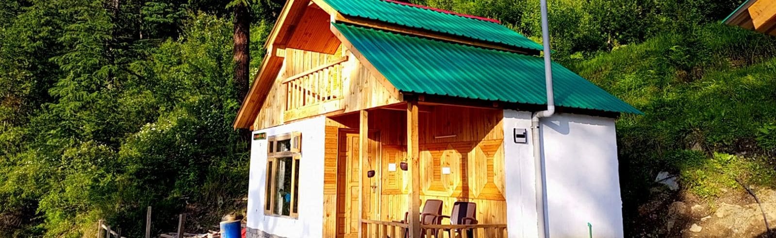 A wooden cabin with a green roof, surrounded by dense forest, and a balcony with chairs.