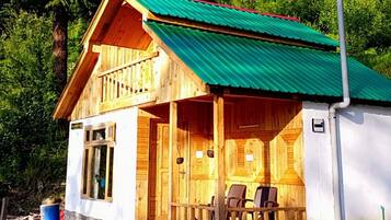 A wooden cabin with a green roof, surrounded by dense forest, and a balcony with chairs.