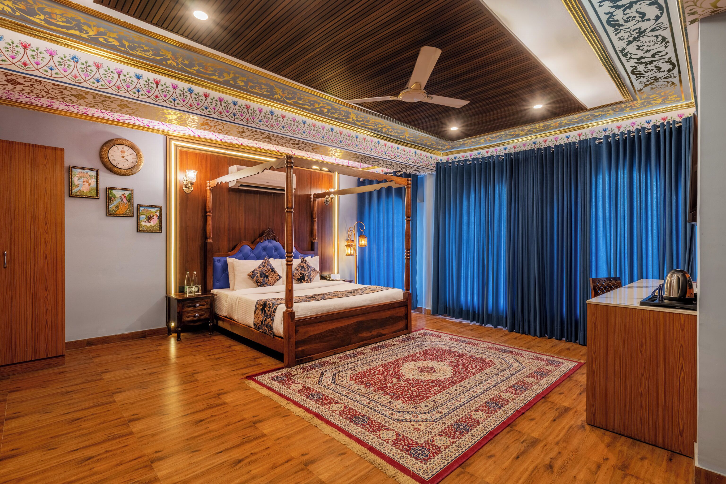 A four-poster bed with a blue headboard, a wooden floor, and a red and beige patterned rug.