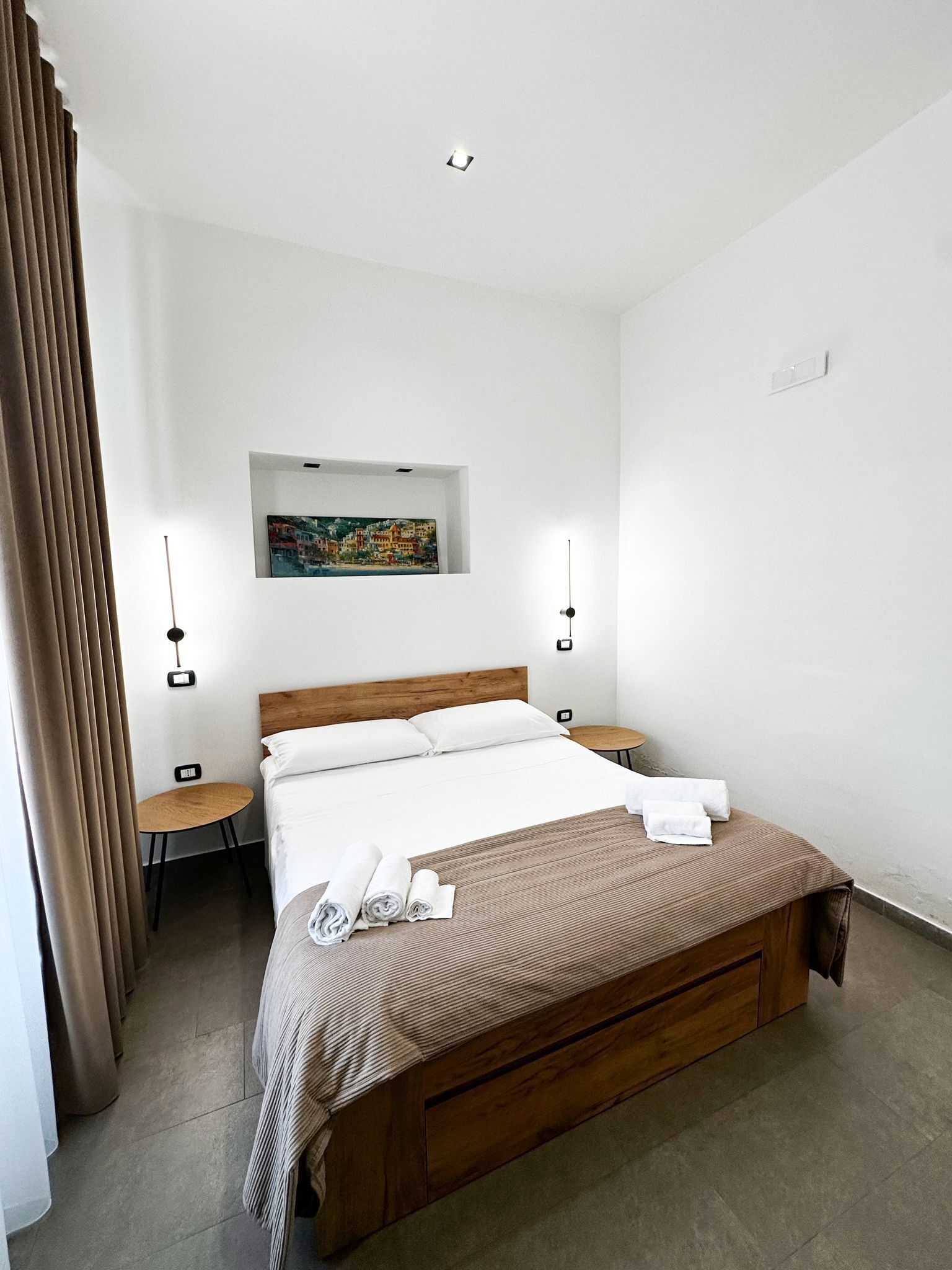 Una habitación de hotel moderna con una cama de madera, una mesita de noche pequeña, una lámpara de noche y un cuadro en la pared, además de una ventana con cortinas.