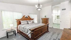 A bedroom with a large bed, a nightstand, a dresser, a chandelier, and a bathroom visible through an archway. - Large Family Home with Game Room (Page)
