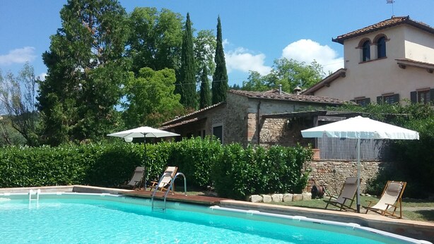 A swimming pool with lounge chairs and umbrellas, surrounded by greenery and a stone building with a tiled roof.