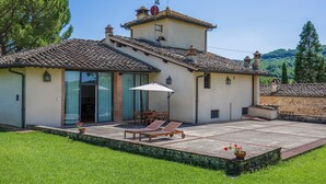 A two-story building with a tiled roof, a patio with a table and chairs, and a grassy lawn.