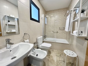 A modern bathroom with a sink, bidet, toilet, and shower. There are shelves with toiletries and a small stool. - Hotel Apartamentos Marina Luz (Chipiona)