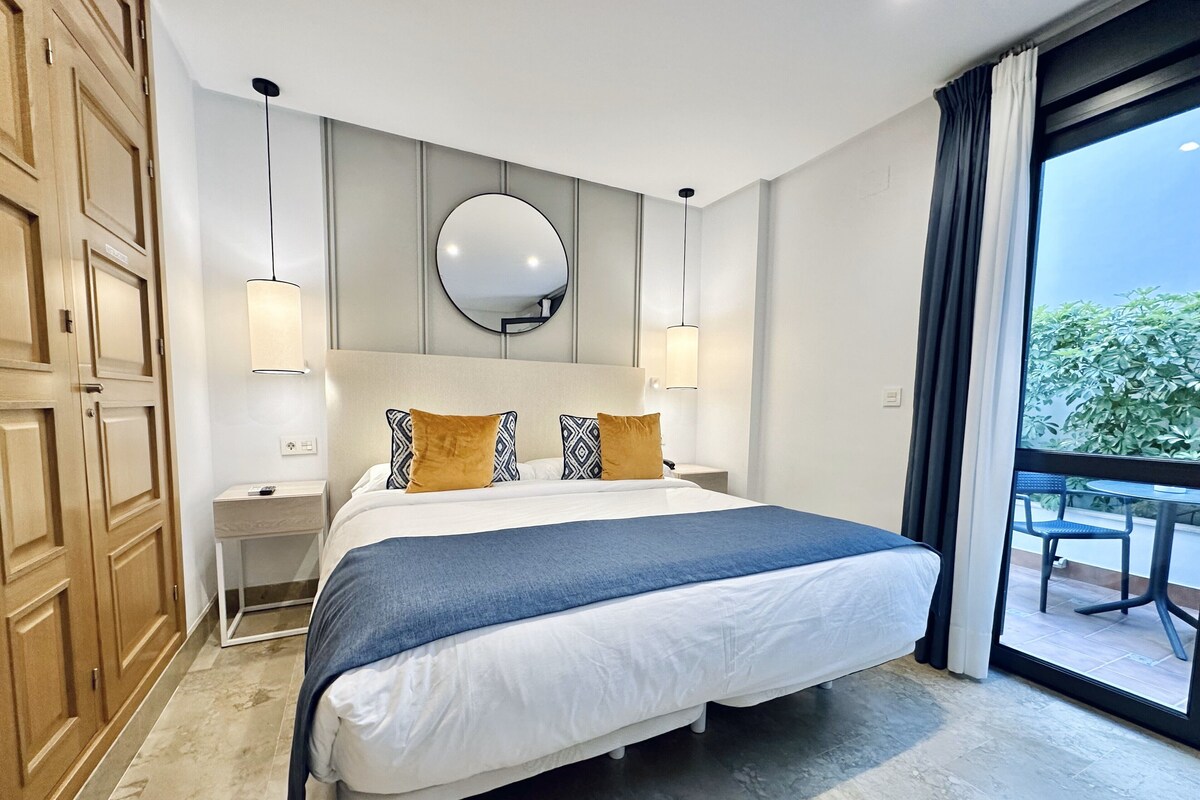 A hotel room with a large bed, a round mirror above the bed, two bedside tables, and a view of greenery through a large window.