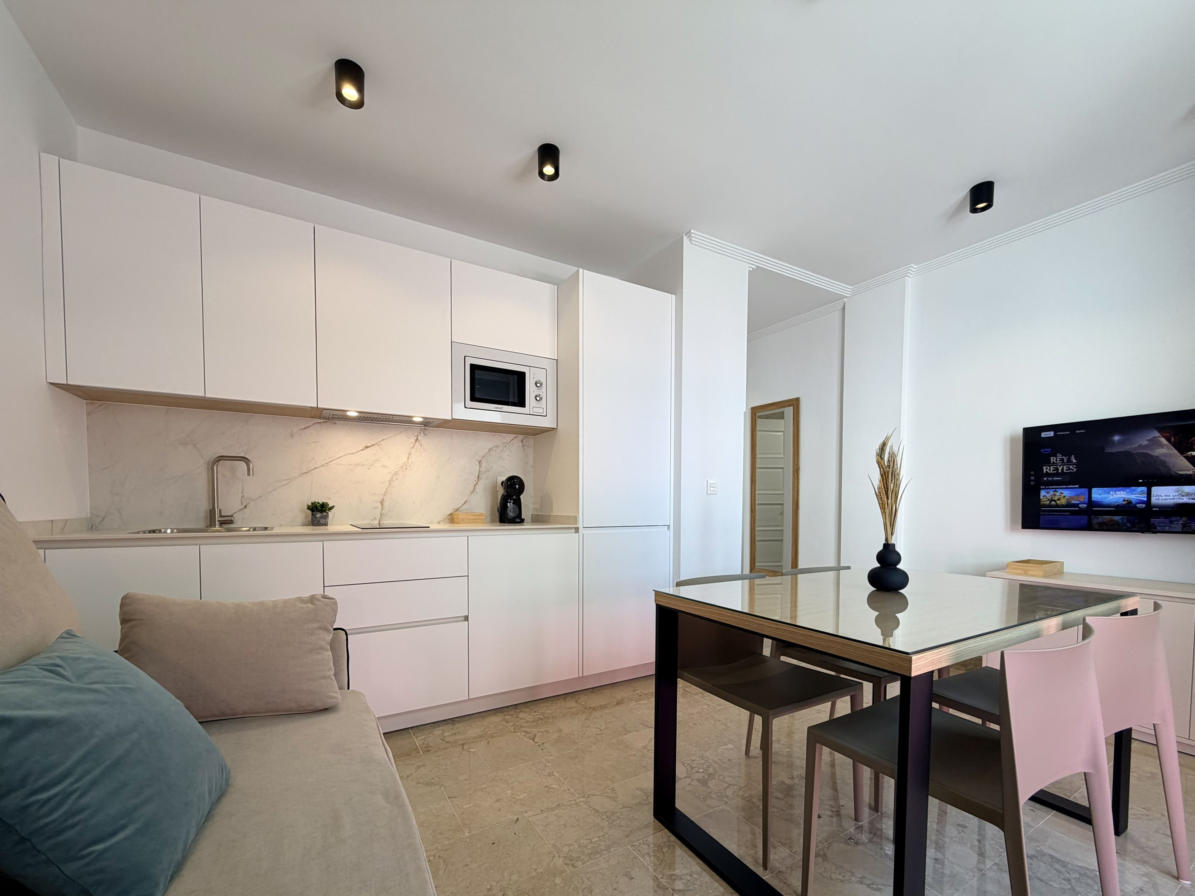 Una cocina moderna con armarios blancos, microondas integrado y un comedor con mesa y sillas de vidrio.