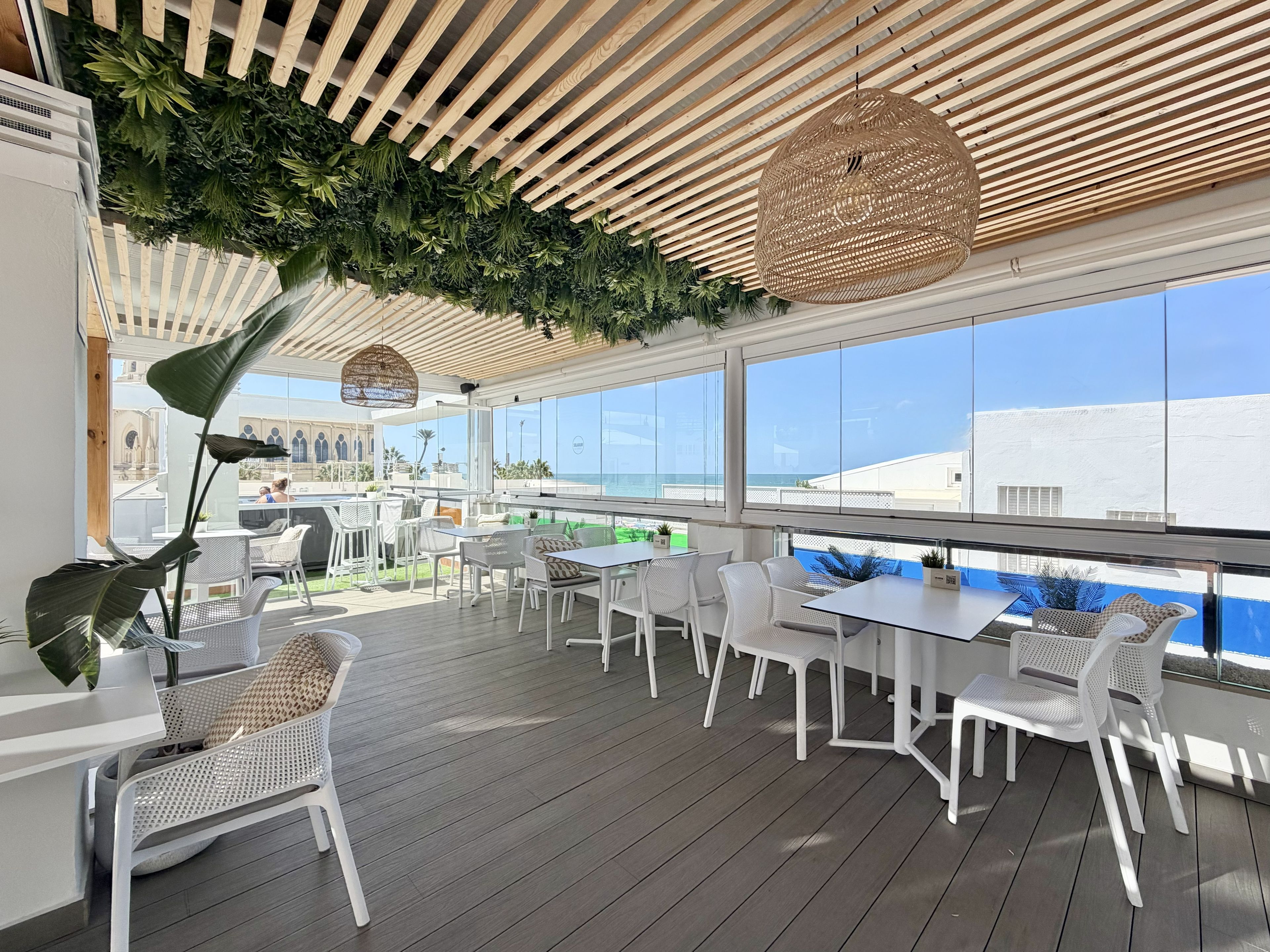 An outdoor seating area with white furniture, a wooden ceiling, and large windows overlooking a cityscape.