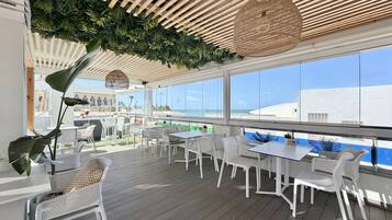 An outdoor seating area with white furniture, a wooden ceiling, and large windows overlooking a cityscape.