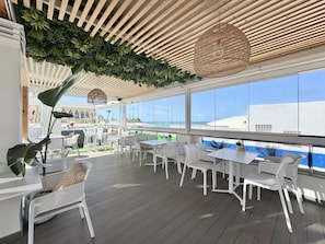 An outdoor seating area with white furniture, a wooden ceiling, and large windows overlooking a cityscape. - Hotel Apartamentos Marina Luz (Chipiona)