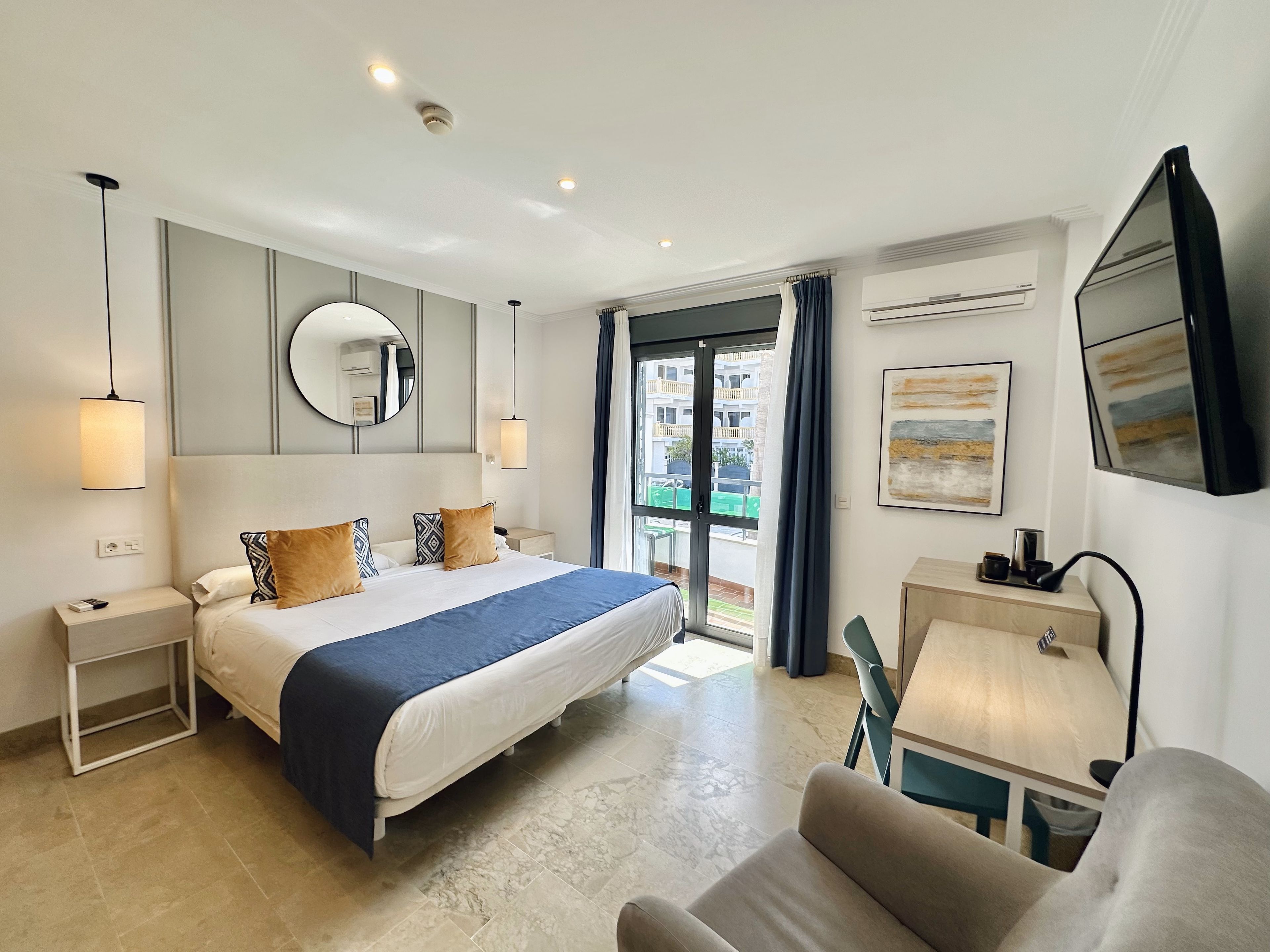 Una habitación de hotel moderna con una cama grande, mesitas de noche, un escritorio y vistas a la ciudad desde la ventana.