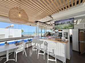 A modern bar area with a large TV screen, white bar stools, and a view of the ocean.