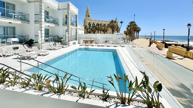 Un área de piscina con sillas de descanso, una piscina azul cristalina y vistas a un edificio histórico con torre.