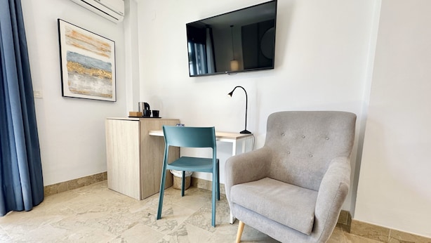 A modern room with a grey armchair, a blue chair, a small desk, a mounted TV, and a painting on the wall. - Hotel Apartamentos Marina Luz (Chipiona)