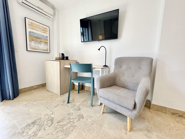 A modern room with a grey armchair, a blue chair, a small desk, a mounted TV, and a painting on the wall. - Hotel Apartamentos Marina Luz (Chipiona)