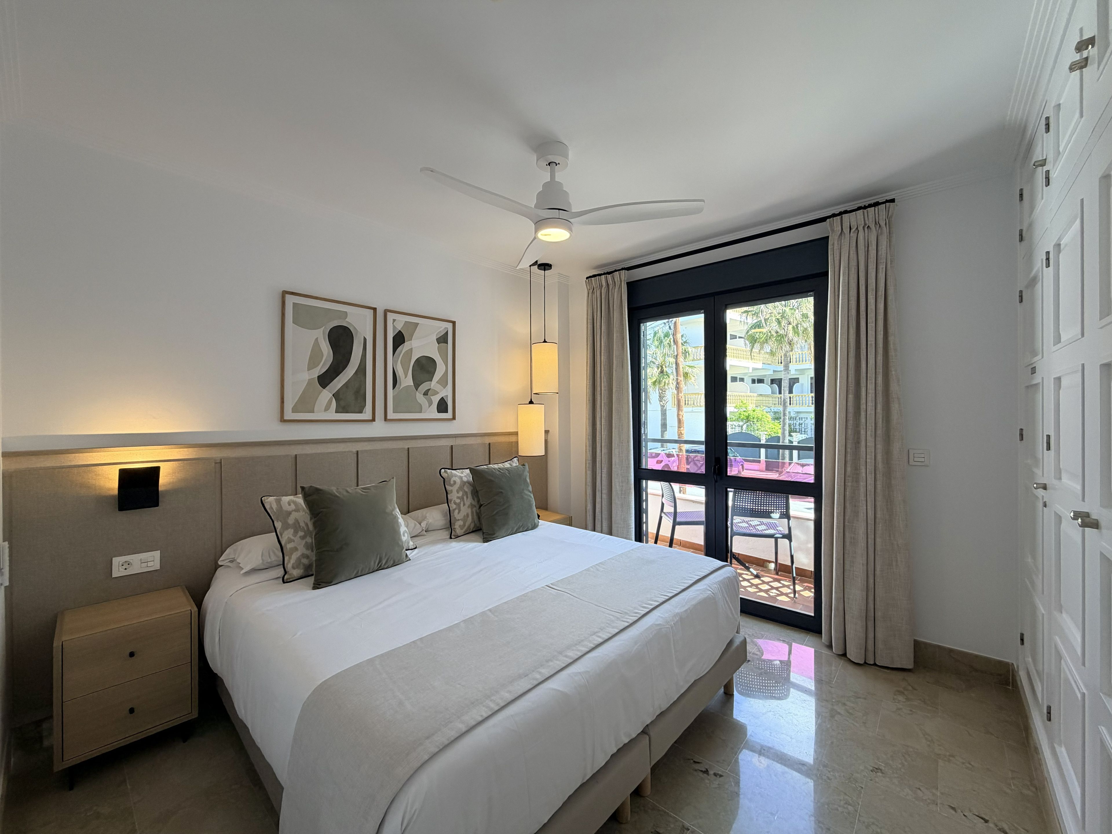 A hotel room with a bed, bedside tables, a ceiling fan, a window with curtains, and artwork on the wall.