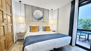 Habitación de hotel con una cama grande, un espejo redondo sobre la cama, dos mesitas de noche y vistas a la vegetación a través de un ventanal amplio.
