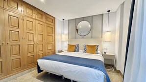 A bedroom with a large bed, wooden wardrobe, bedside lamps, and a round mirror on the wall. - Hotel Apartamentos Marina Luz (Chipiona)