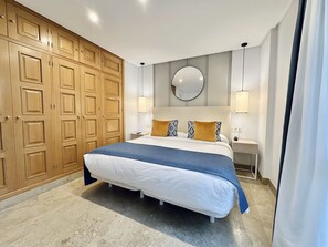 A bedroom with a large bed, wooden wardrobe, bedside lamps, and a round mirror on the wall. - Hotel Apartamentos Marina Luz (Chipiona)
