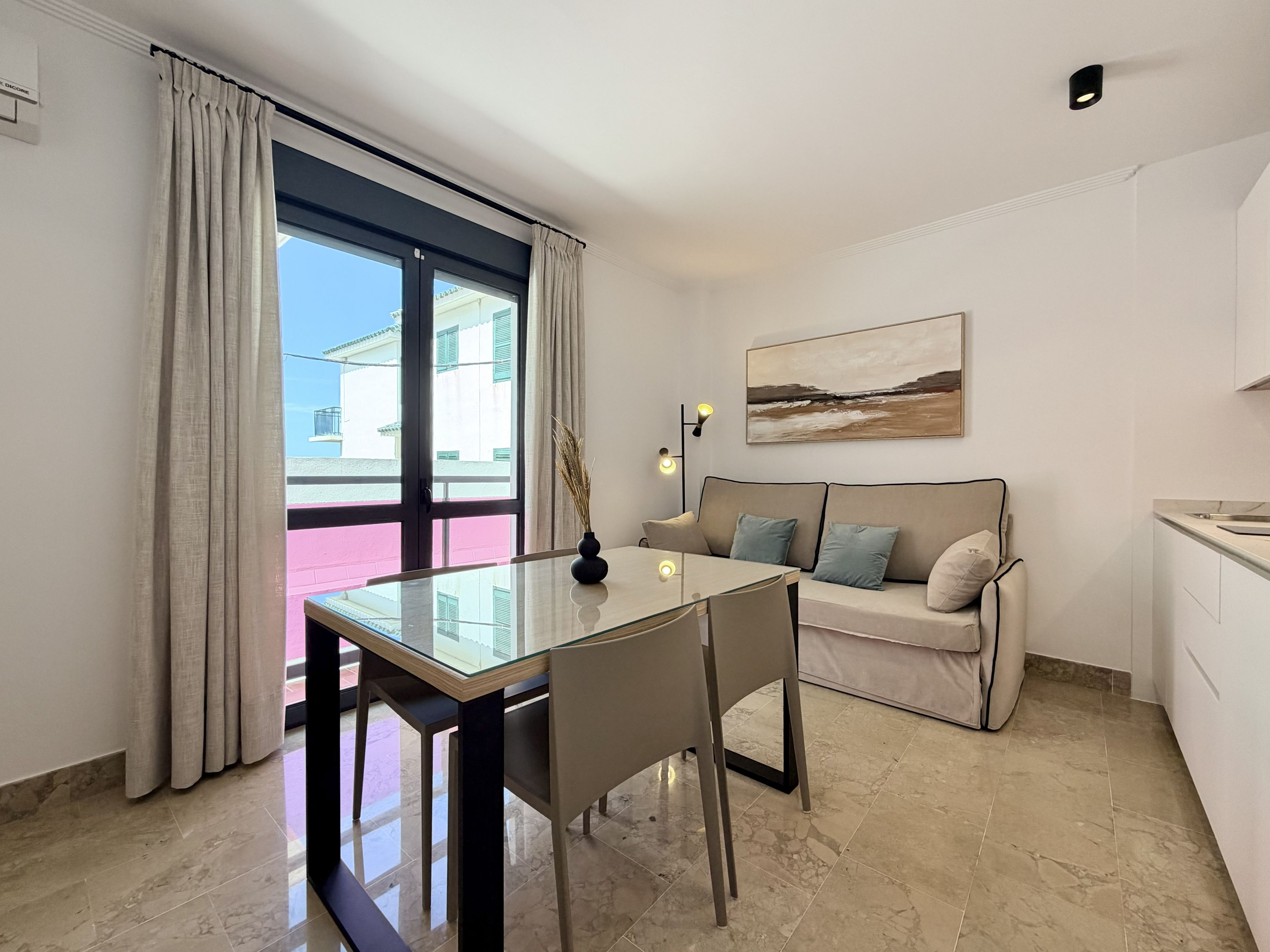 Un salon moderne avec une table à manger en verre, un canapé avec des coussins, un tableau au mur et une vue à travers une grande fenêtre.
