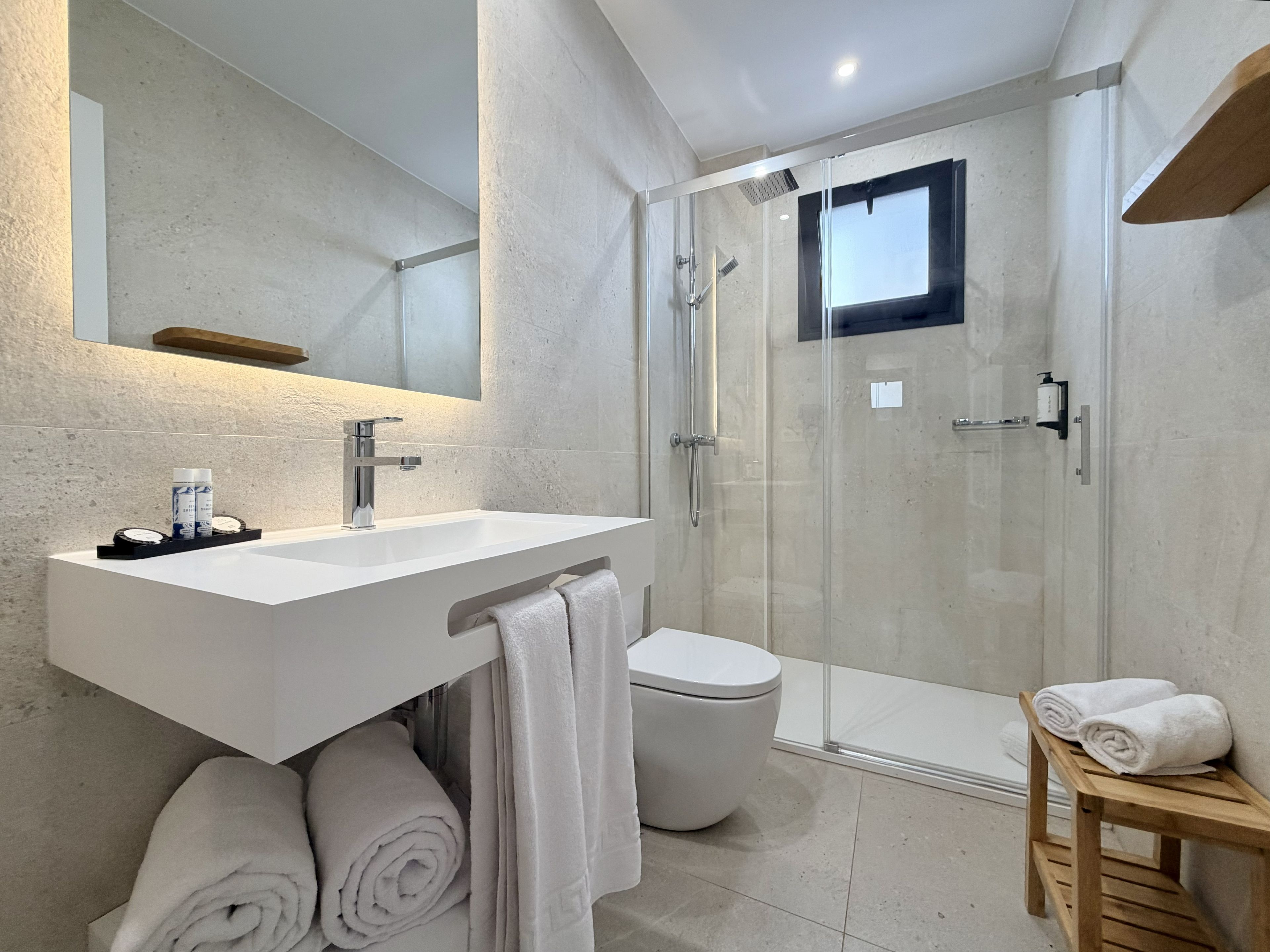 Un baño moderno con un lavamanos blanco, cabina de ducha de vidrio, inodoro y un banco de madera con toallas.