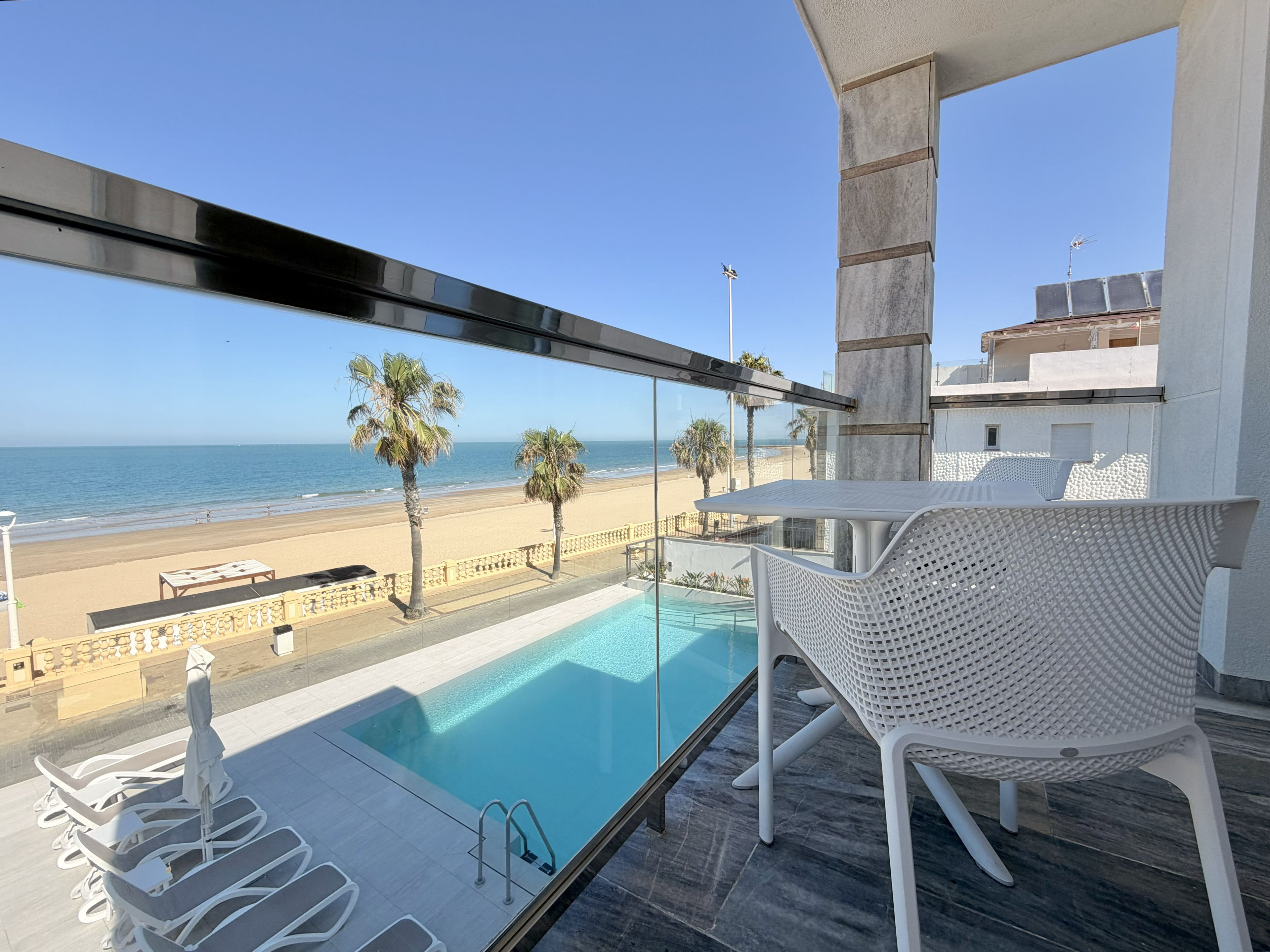 Un balcón con piscina, sillas blancas y vista a la playa y al océano.