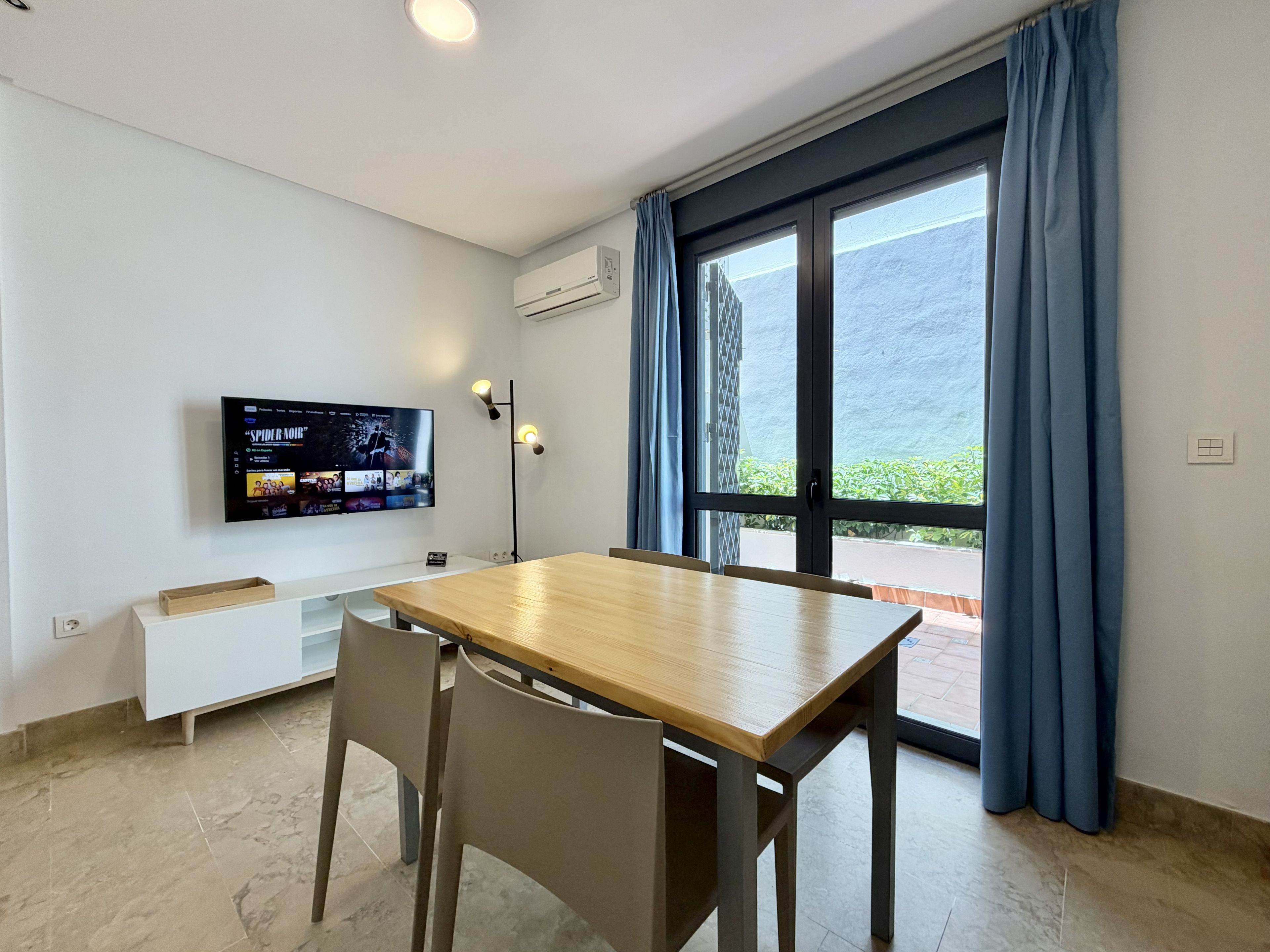 Una habitación con una mesa de madera, dos sillas, un televisor montado en la pared y una ventana con cortinas.