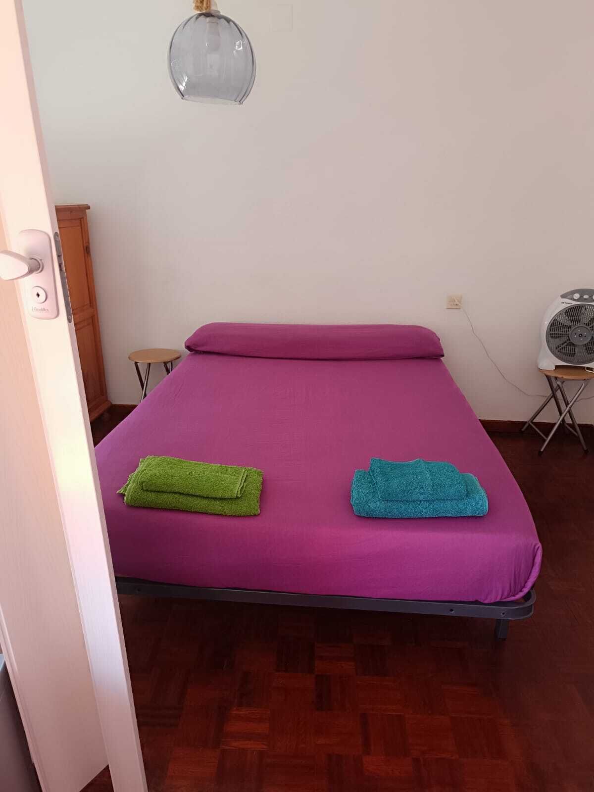 Una habitació amb un llit, una taula petita, un ventilador i una porta.