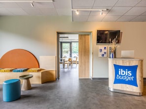 A modern interior with a reception desk labeled 'ibis budget,' a seating area with a sofa and ottoman, and a view into a dining area.