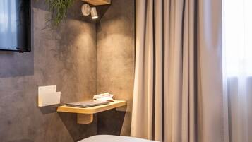 A modern hotel room with a bed, a mounted TV, a bedside table with a laptop, a wall-mounted light, and a curtain.