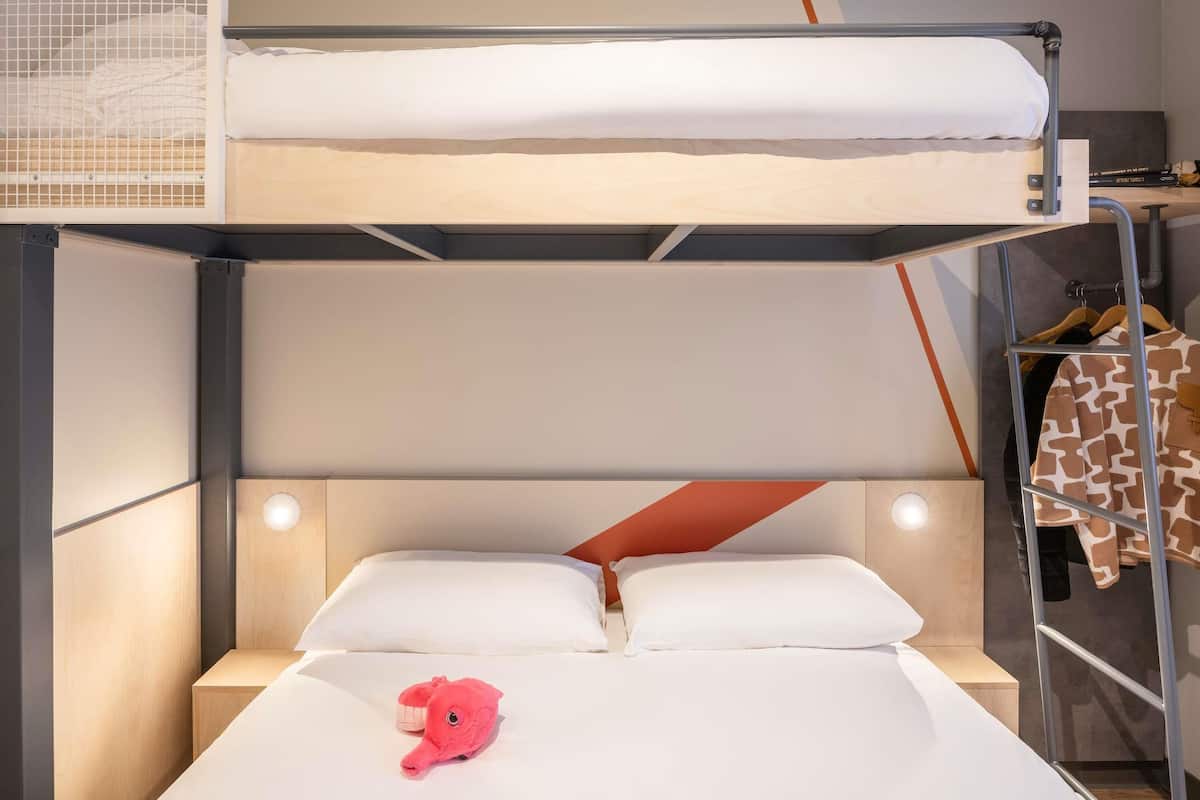 Un lit superposé avec un lit en bas et un lit en haut. Il y a une petite étagère avec une lampe, et une veste à motifs de girafe est accrochée à un porte-manteau.