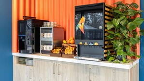 A coffee station with a coffee machine, a fruit display, and a laptop on a counter against an orange wall. - ibis budget Arlon Porte du Luxembourg (Arlon)