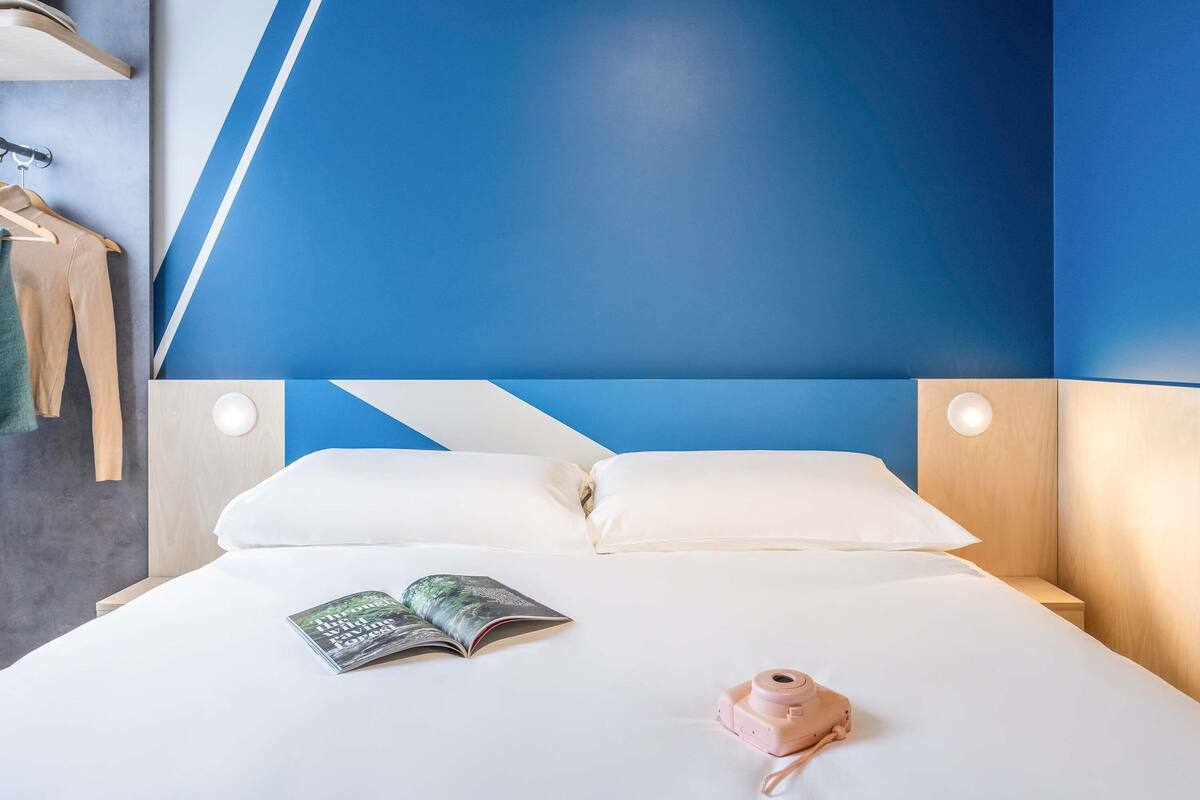 Un lit bien fait, avec des draps blancs, un appareil photo rose et un livre ouvert posé sur le lit.