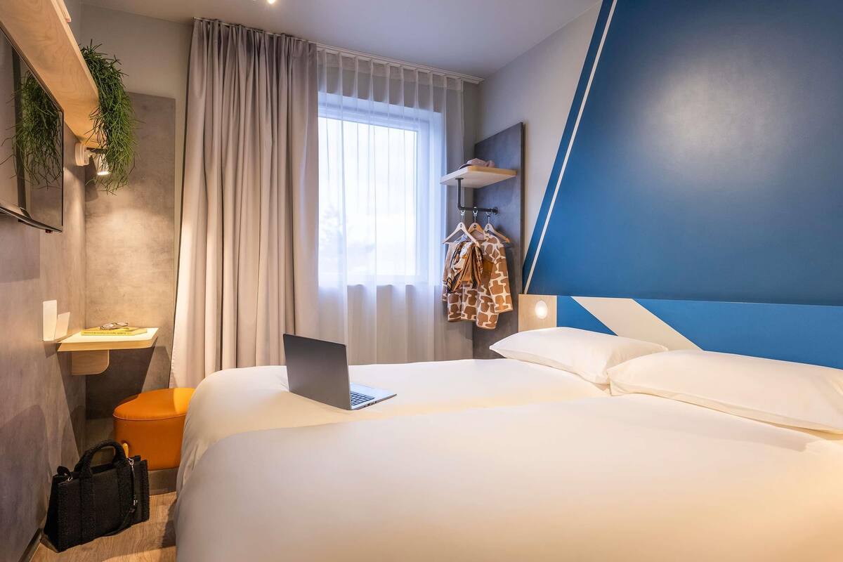 Une chambre d’hôtel avec un lit, un bureau avec un ordinateur portable, un porte-manteau et une fenêtre avec des rideaux.