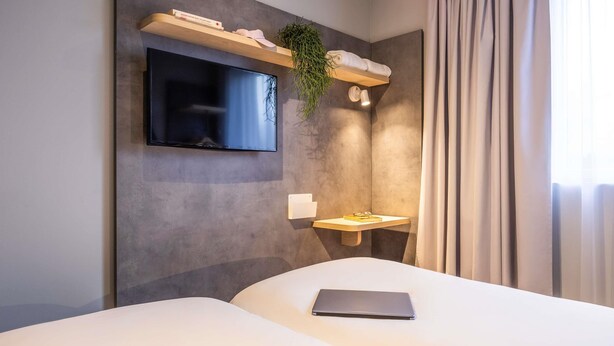 A modern hotel room with a flat-screen TV mounted on the wall, a bedside table with a laptop, a small shelf with a plant and a towel, and a window with curtains. - ibis budget Arlon Porte du Luxembourg (Arlon)