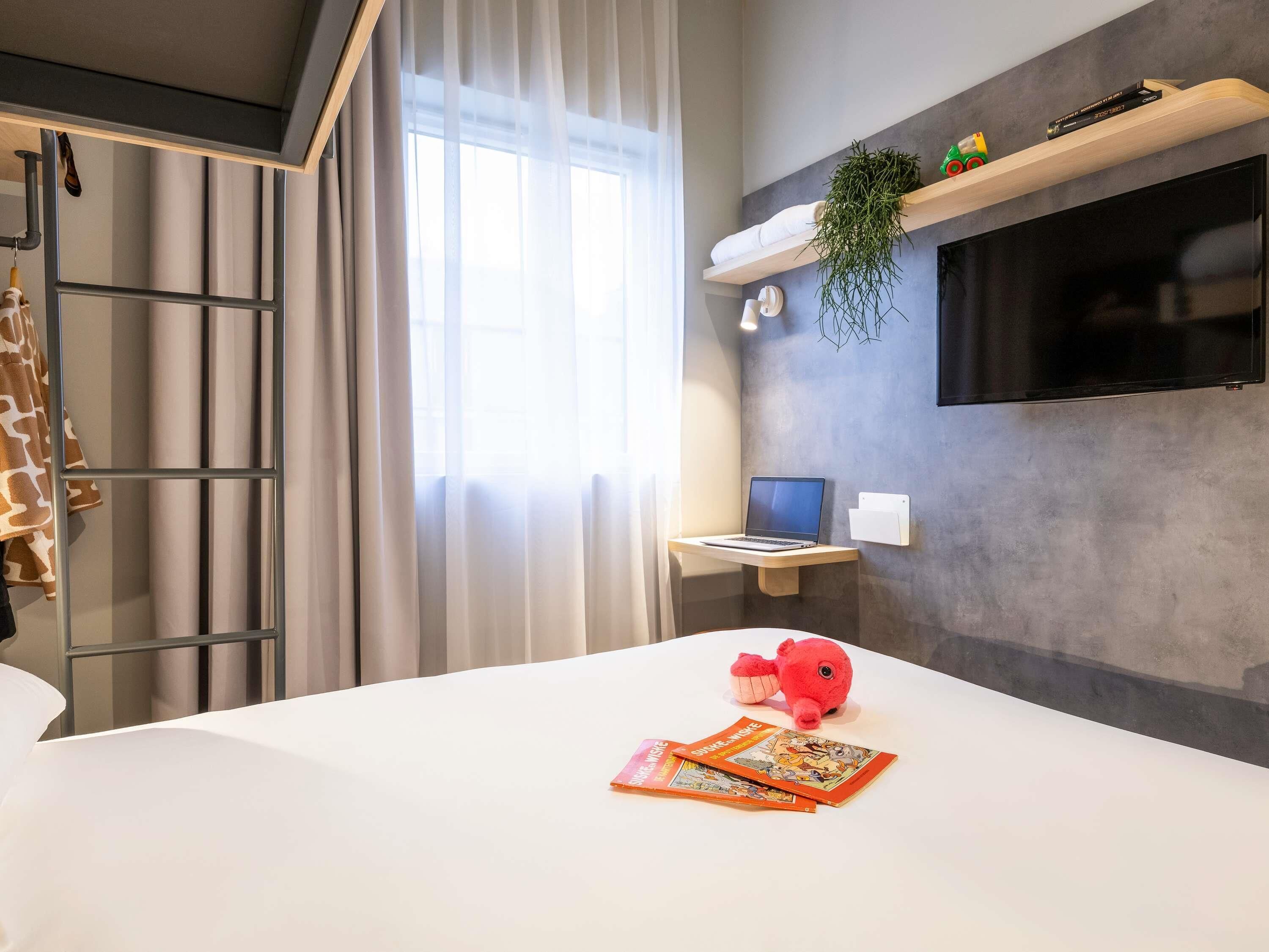 Una habitación de hotel moderna con una cama, un escritorio con una computadora portátil, un televisor montado en la pared y una ventana con cortinas.