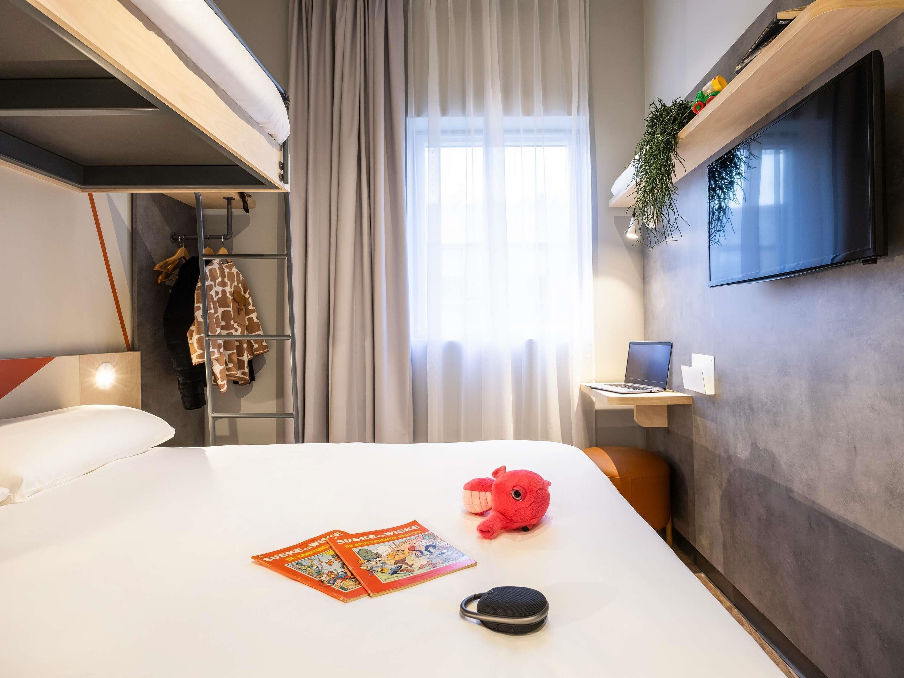A hotel room with a bunk bed, a desk with a laptop, a television, a plant, and a stuffed toy on the bed.