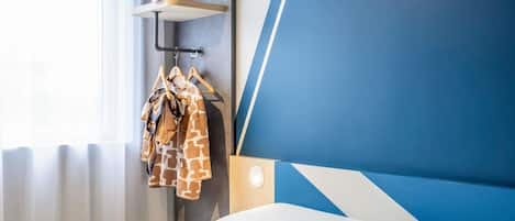 Una habitación de hotel con una cama, cabecero, perchero en la pared con una chaqueta de estampado de jirafa y una ventana con cortinas.